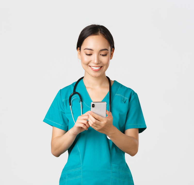 Covid-19, healthcare workers and preventing virus concept. Smiling happy beautiful asian female intern, doctor making phone call, looking at mobile phone screen pleased, messaging or using app